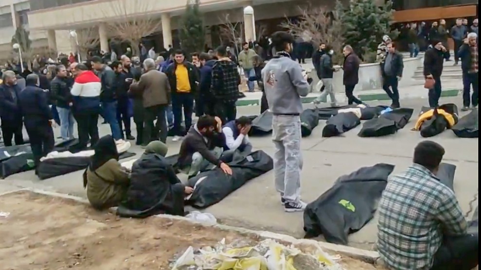 Dozens of people crowd around bodies in black bags. Some are kneeling or sitting while others are standing. One man in the center has his hands covering his eyes.